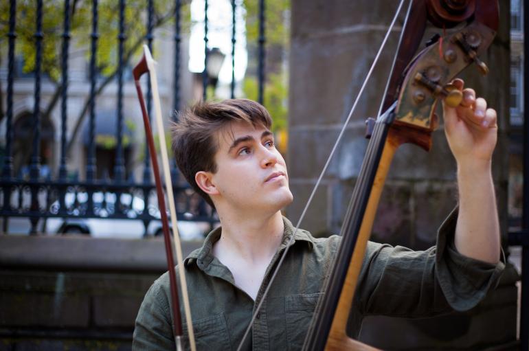 Bass player Sam Suggs, with messy hair, looks up at the peg box of his bass as he loosens a string several inches