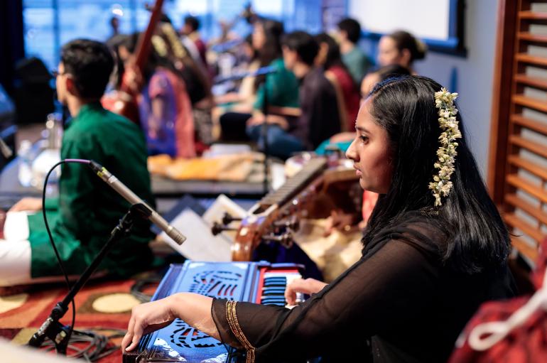 A young woman sits at a South Asian instrument wearing colorful clothing