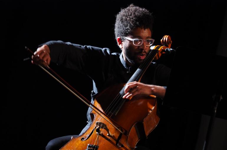 Seth Parker Woods wearing black and playing cello in a dark room, lit from the side