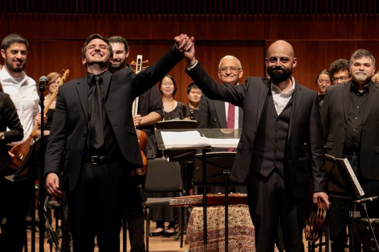 Directors of the chamber orchestra and middle east music ensemble stand smiling in front of an orchestra with their hands clasped together