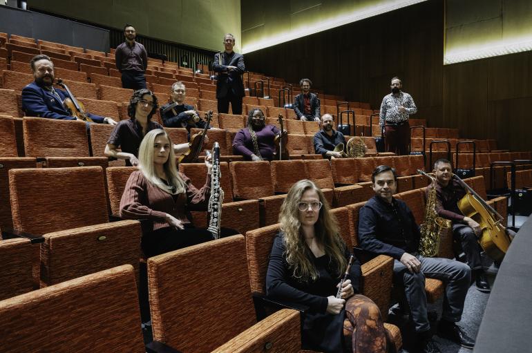A group of musicians sits in the seats of a performance hall. They are sitting spaced out in different rows and seats, staring powerfully at the camera with their instruments.