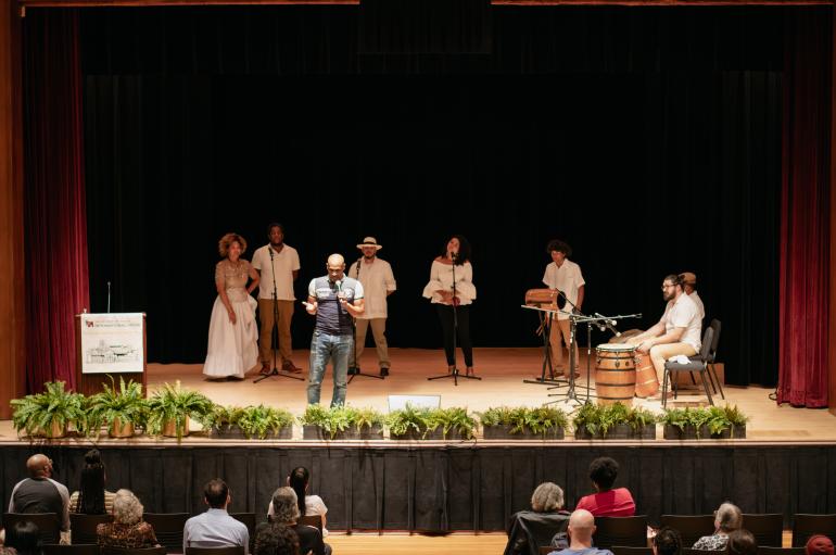 A man stands on stage speaking into a microphone, with a small group of musicians with drums and microphones behind him