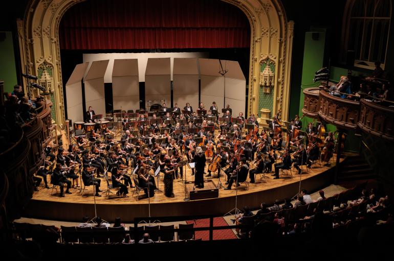 Around 100 instrumentalists dressed in black on stage performing music with a conductor in an ornate concert hall