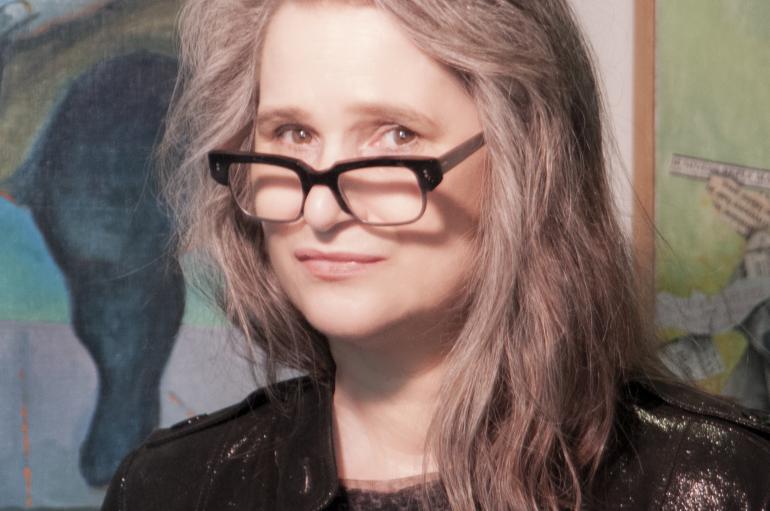 Headshot of a woman with long gray hair and glasses standing in front of a painting. Credit Valerie Booth