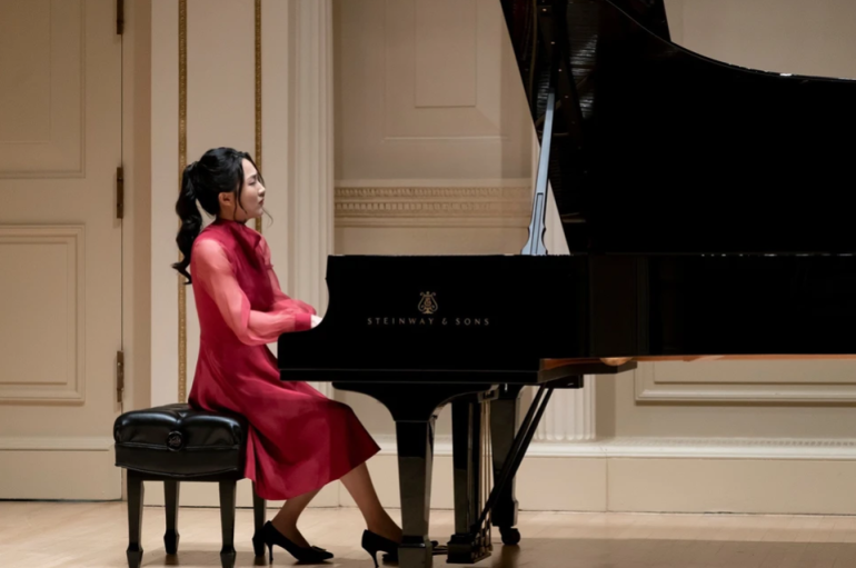 A woman in a red long sleeved dress closes her eyes as she plays the piano