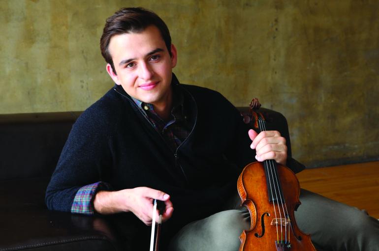 A man in a navy collared sweater leans casually on a couch while holding a violin