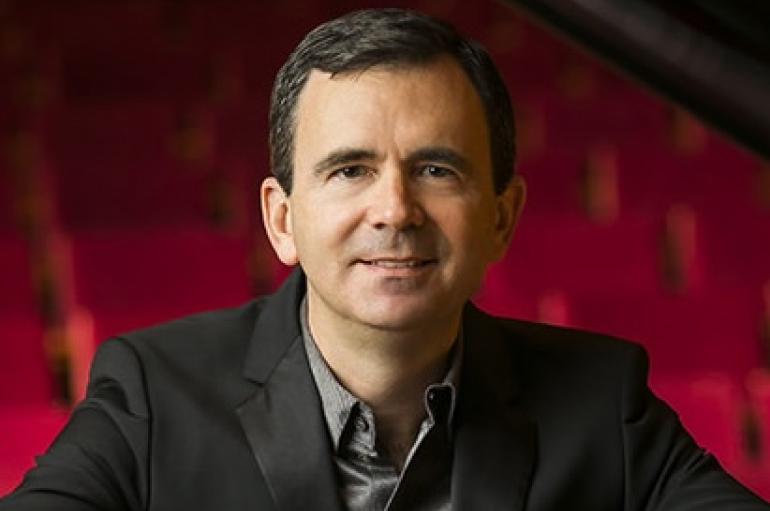 A headshot of a man in a dark suit smiling in front of a red background