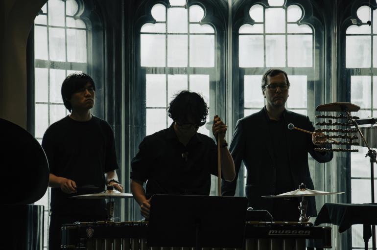 Three percussionists perform, silhouetted in front of ornate windows