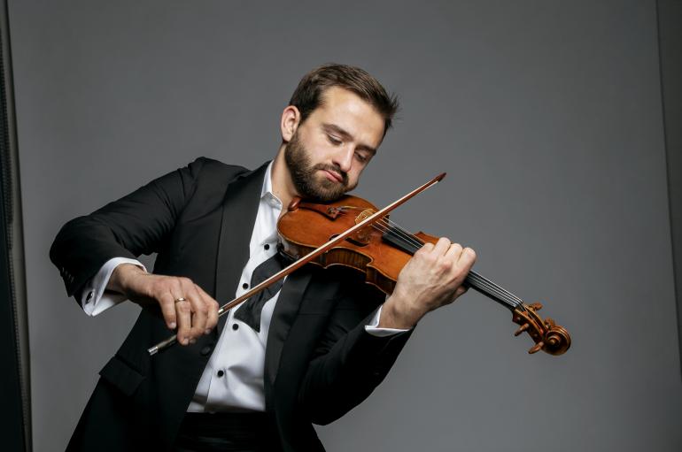 A man in a tuxedo plays the violin in front of dark gray backdrop