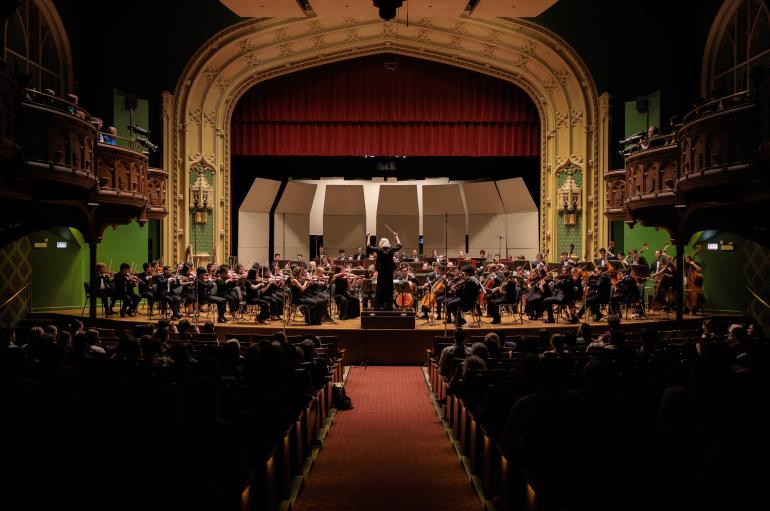 A view from the back of a concert hall. An orchestra is performing on a stage with green, gold, and red detailing.