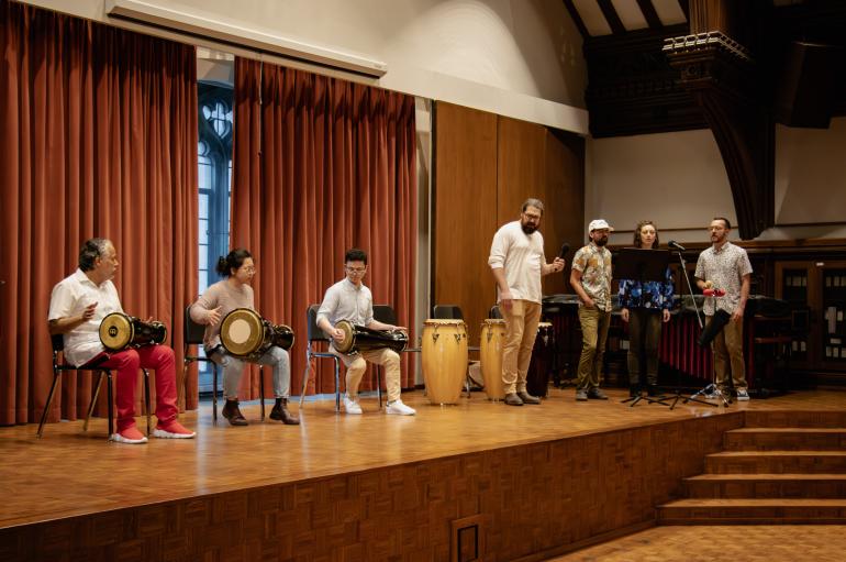 Side angle view of a concert stage. Three musicians are seated and playing on traditional drums, and three stand off to the side signing into a microphone