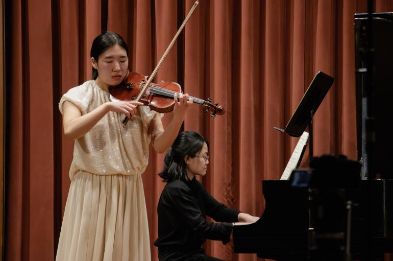A girl in a beige dress plays a violin with her eyes closed, accompanied by a pianist sitting behind her. They are on a stage in front of a red curtain.