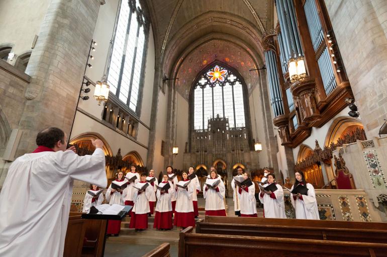 A man in white robes conducts a small group of singers in white robes in an ornate chapel