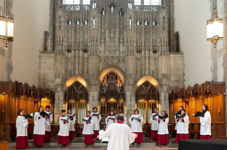 A man in white robes conducts a small group of singers in white robes in an ornate chapel