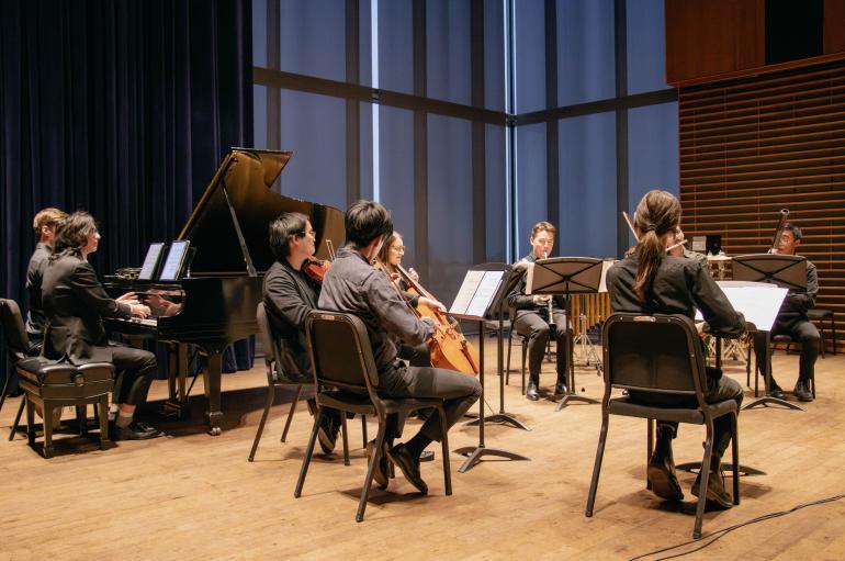 Side view of a small group of musicians performing on stage. They are seated and have music stands in front of them.