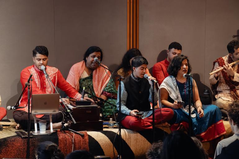 A group of musicians are seated on the floor, wearing traditional South Asian clothing, and singing into microphones