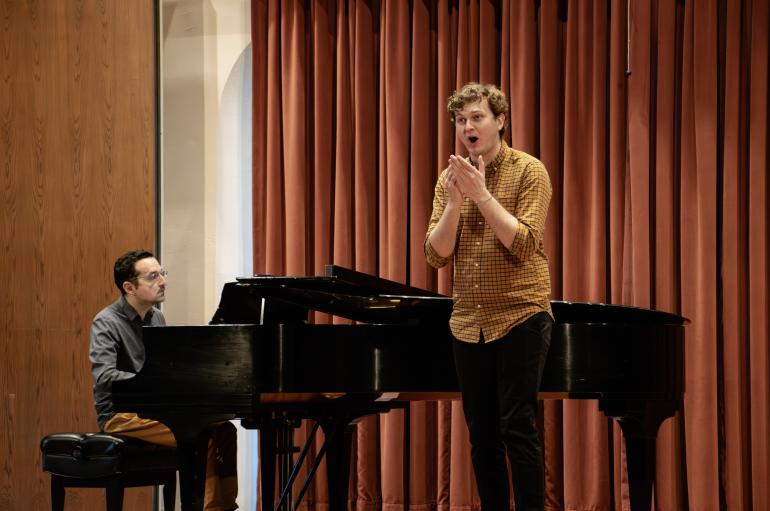 A male singer performs on stage, with a piano accompanist sitting behind him