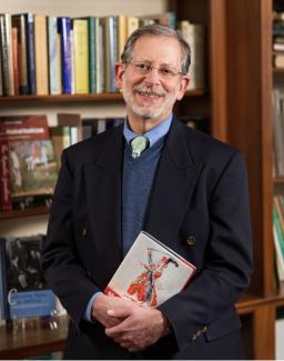 Ralph P. Locke stands wearing a navy sport coat, blue sweater, light blue shirt, and green and blue paisley tie holding one of his books