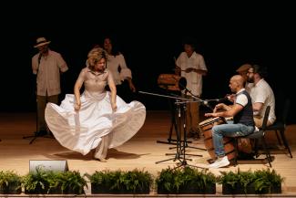 A woman with a long white skirt dances on stage next to a small group of musicians playing traditional drums