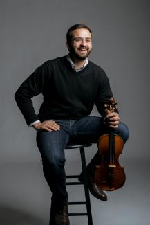 A man sits on a stool in front of a gray background. He smiles into the distance, holding a violin in his left hand.