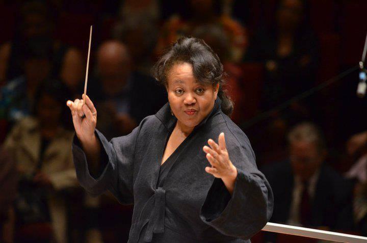 Renée Baker, dressed in black, stands on the conductors podium, arms outstretched, holding a baton, with audience in the background