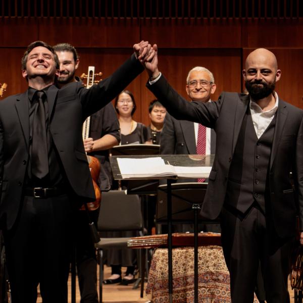 Directors of the chamber orchestra and middle east music ensemble stand smiling in front of an orchestra with their hands clasped together