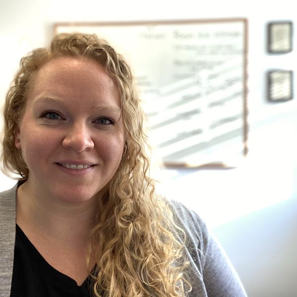 A woman with blonde curly hair smiles in front of a sunlit wall