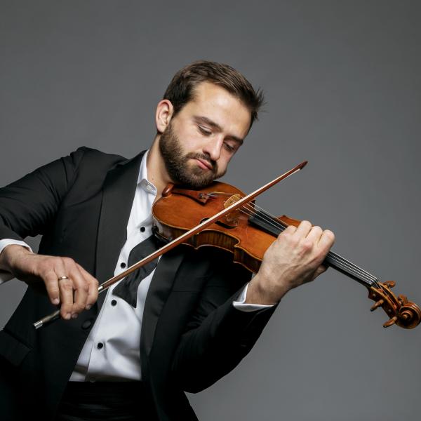 A man in a tuxedo plays the violin in front of dark gray backdrop