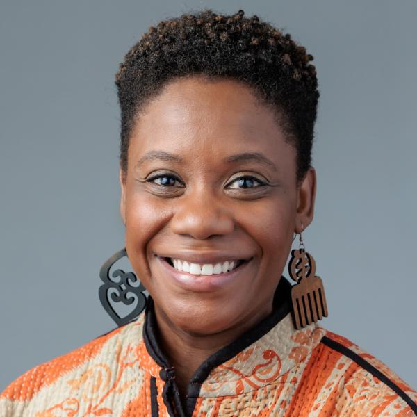 Headshot photo of Jessica Baker in front of grey background. She is wearing an orange patterned top and wood cut earrings.